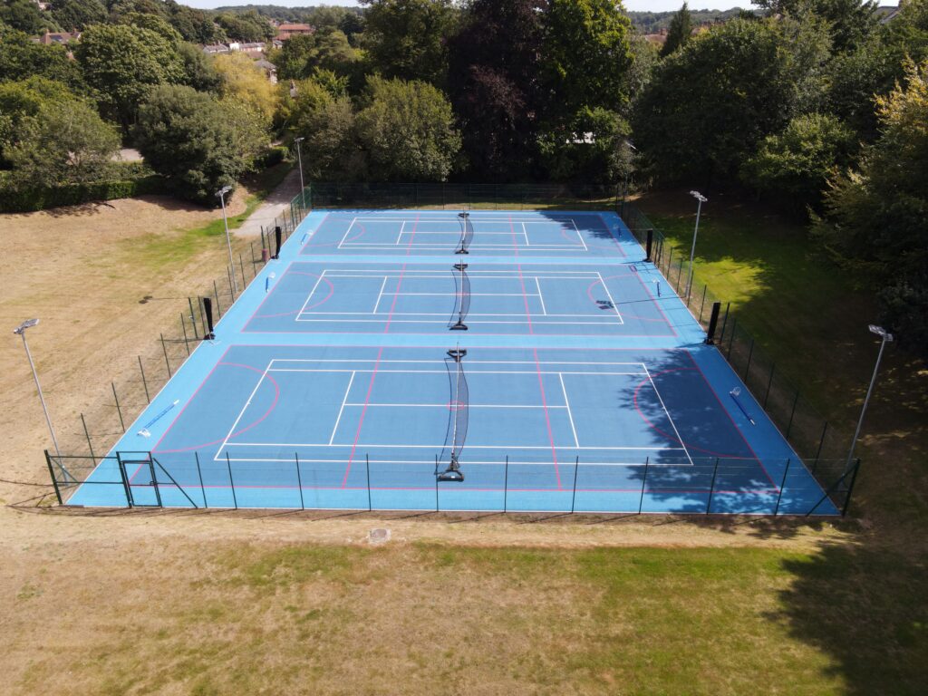 3 blue netball courts with fence around and floodlights. Area surrounded by grass and trees.